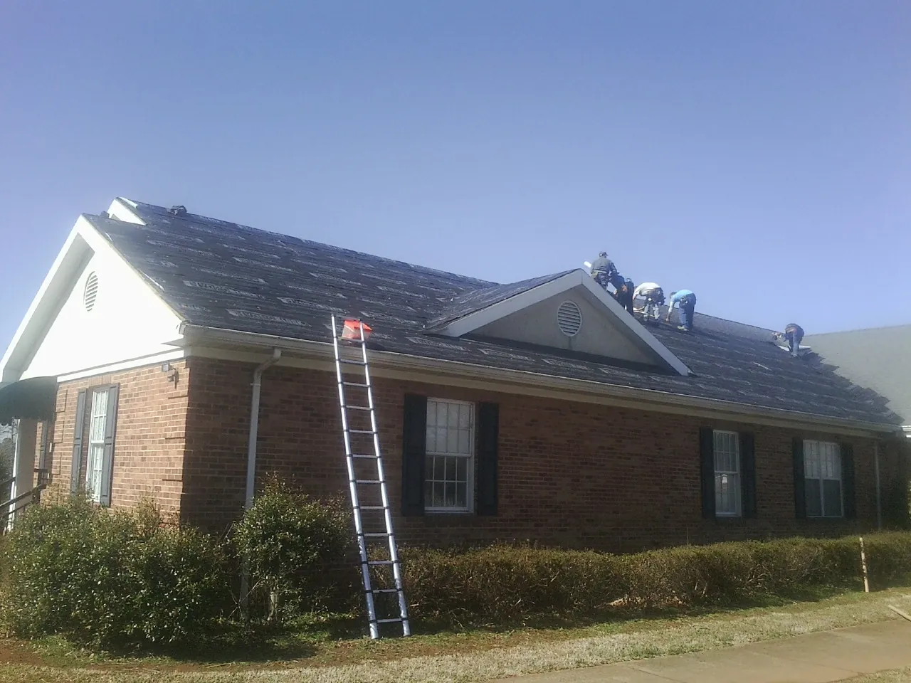 Crew tearing shingles off a brick ranch roof with a ladder set — Emergency Roof Repair work by our crew in Luke