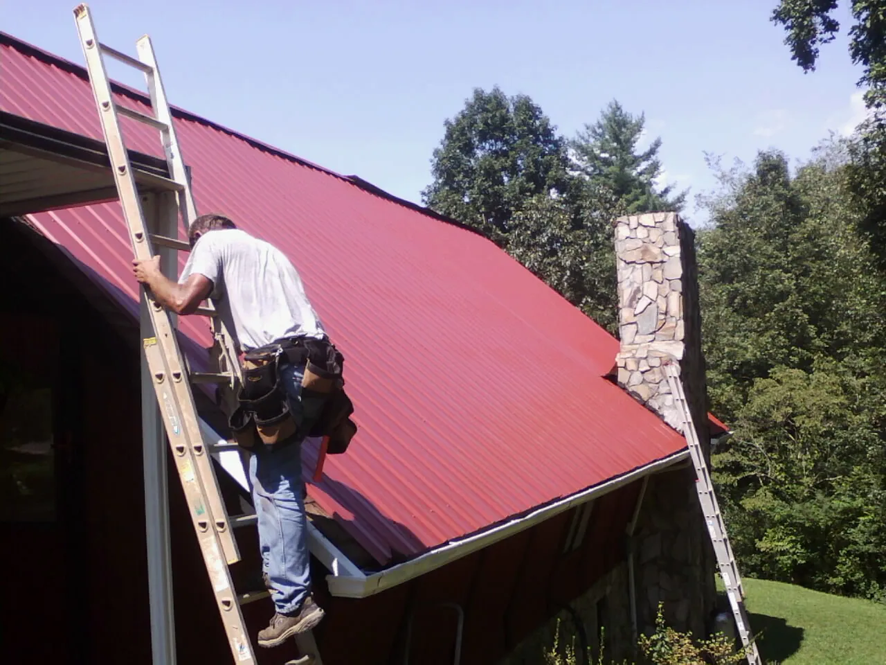 Roofer on a ladder beside a red metal roof and stone chimney — Soffit & Fascia Repair by Roofing Brothers in Luke
